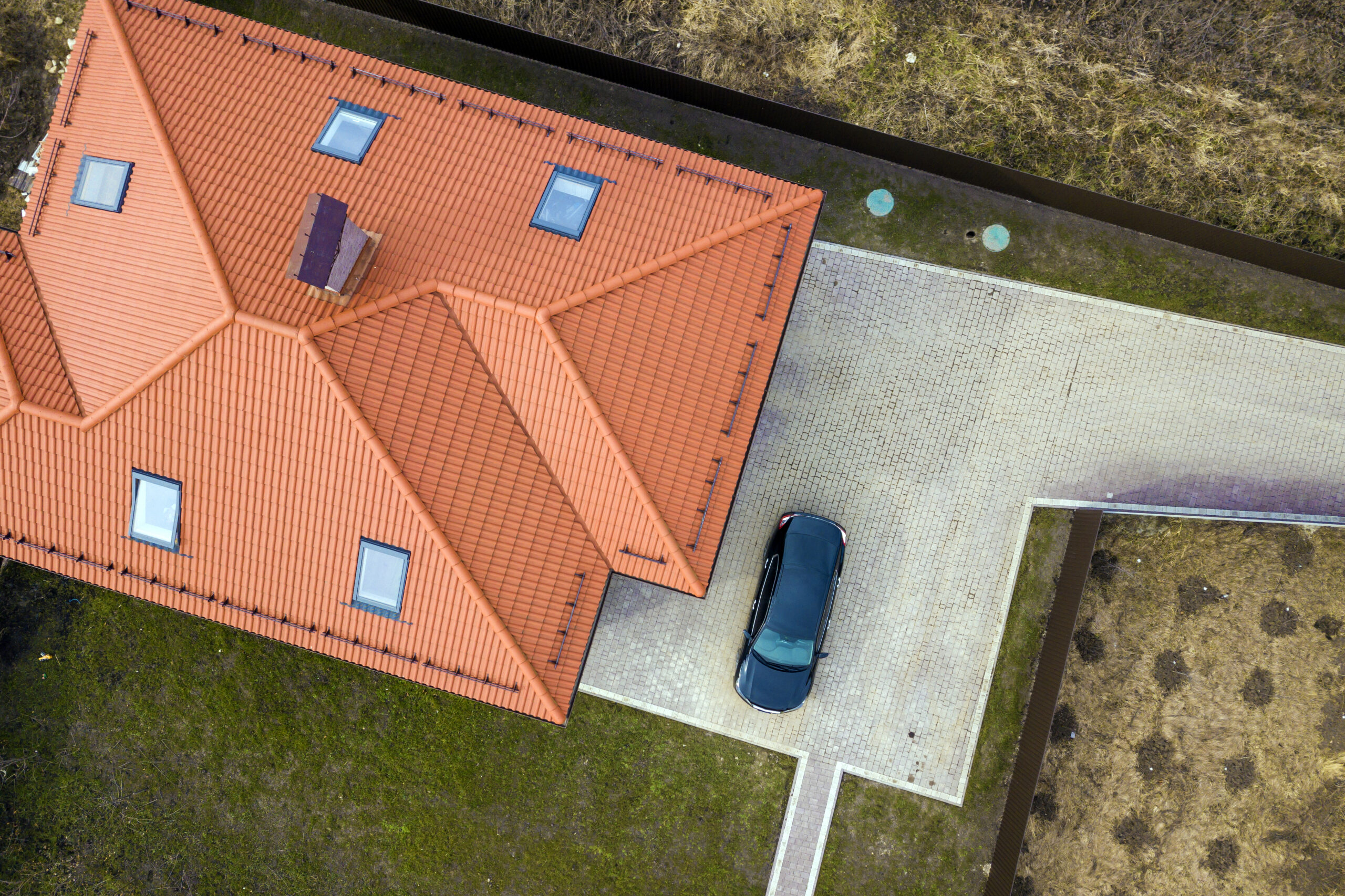 Aerial top view of house metal shingle roof with attic windows and black car on paved yard.